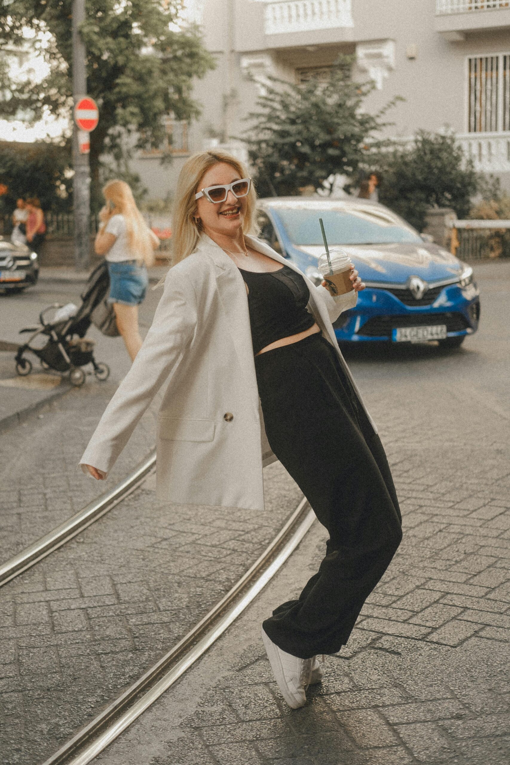 Stylish woman in urban setting wearing black crop top and white blazer while posing on tram tracks.